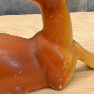 May include: A ceramic deer figurine in a lying down position. The deer is brown and yellow with a textured surface.