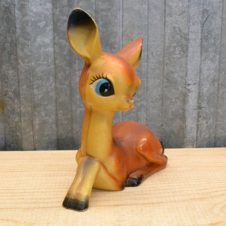 May include: A vintage ceramic deer figurine with large blue eyes and a brown and yellow painted body. The deer is lying down with its head turned to the side.