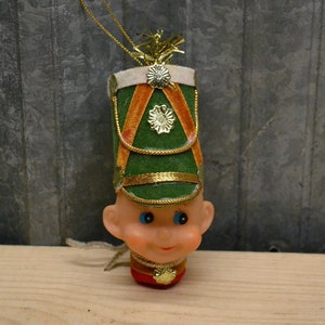 May include: A vintage Christmas ornament shaped like a toy soldier's head. The ornament is made of plastic and has a green hat with gold trim, a white face with blue eyes, and a red and gold uniform.