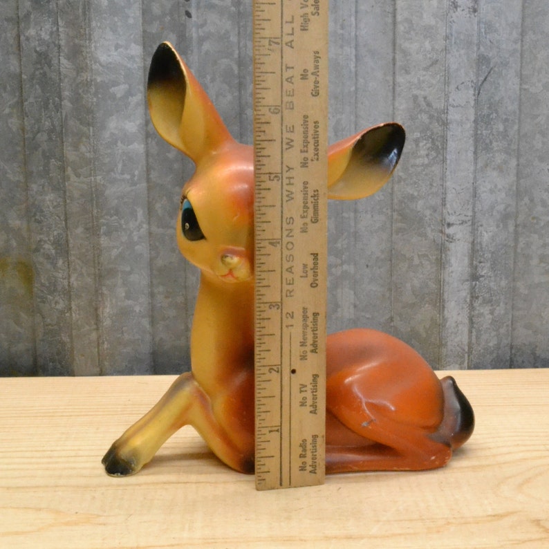 May include: A brown and orange ceramic deer figurine with blue eyes. The deer is sitting on its hind legs with its front legs outstretched. The figurine is approximately 7 inches tall.
