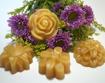以下が含まれることがあります： バラや他の花の形をした、手作りの薄茶色のキャンドルのコレクション。紫色の菊と緑の葉で飾られています。