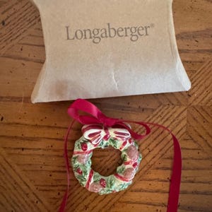 Peut inclure: Une décoration de couronne de Noël en céramique avec un feuillage vert, des baies rouges et un nœud rouge, accompagnée d'un ruban rouge pour la suspendre. L'ornement est présenté avec une boîte en carton beige portant le nom Longaberger en brun.