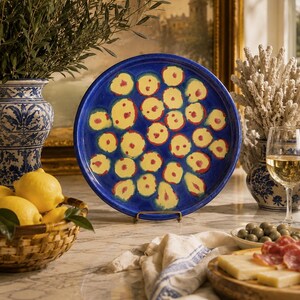 May include: A round ceramic plate featuring a deep blue background decorated with numerous yellow circular shapes, each containing a small red centre. The plate is displayed upright on a small metal stand, showcasing its vibrant, hand-painted pattern.