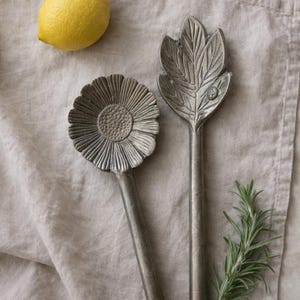 May include: Two decorative serving spoons with floral and leaf designs. One spoon features a sunflower head, the other a leaf. The spoons are silver-colored and rest on a neutral-toned cloth. A yellow lemon and sprig of rosemary are also present.