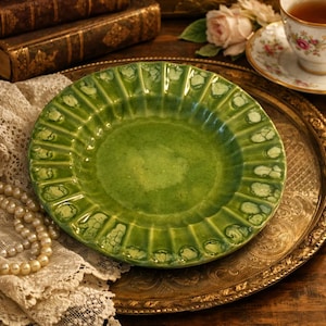 May include: A green ceramic plate with a fluted rim and a textured surface. The plate is set on a gold-toned metal tray, with a pearl necklace and lace in the foreground. A teacup and books are in the background.