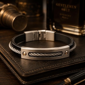 Può includere: Un bracciale con cinturino in gomma nera e una placca centrale in metallo argentato spazzolato. La placca è intarsiata con un design a cavo d'acciaio intrecciato e fissata da due piccole viti dorate su entrambi i lati. Include una chiusura argentata.