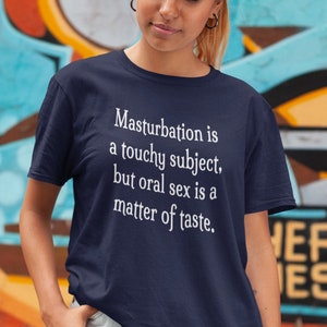 May include: Navy blue t-shirt with the text "Masturbation is a touchy subject, but oral sex is a matter of taste" in white. The shirt is being worn in front of a colorful graffiti background.