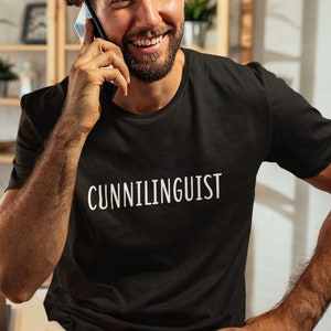 May include: A man wearing a black t-shirt with the word "CUNNILINGUIST" printed in white on the front. He is talking on a cell phone and smiling.
