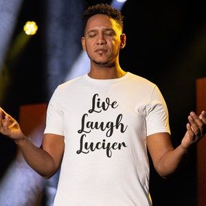 May include: A white t-shirt with the text "Live Laugh Lucifer" in a black script font. The shirt is being worn by a person with their eyes closed and arms raised.