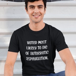 May include: Black t-shirt with the text "Voted Most Likely to Die of Autoerotic Asphyxiation" in white. The shirt is worn by a person squatting down, holding a blue leash, with a brown dachshund dog wearing a harness.
