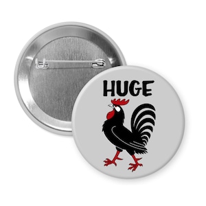 May include: A round button with a gray background and a black rooster illustration. The word "HUGE" is printed in black above the rooster. The rooster has red accents on its comb, wattle, and feet. A second button is visible in the background.