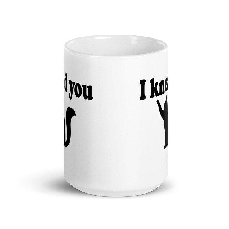 15 ounce white ceramic coffee mug with a pun image of a silhouette of a cat and the words I knead you printed on both sides of the mug.