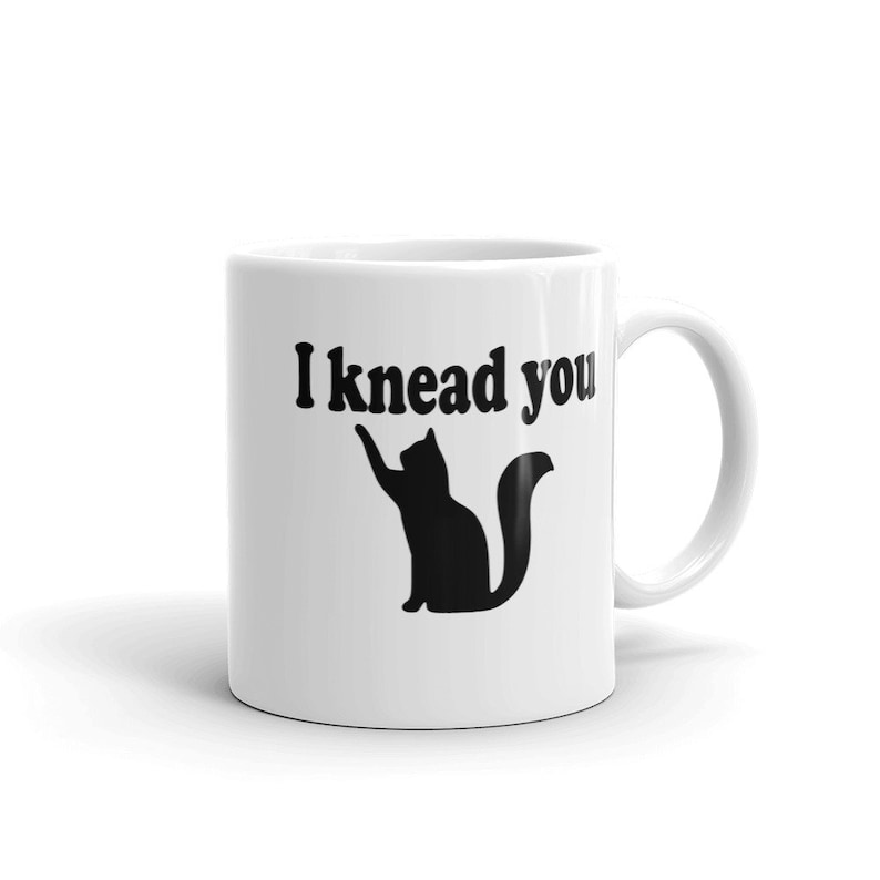 11 ounce white ceramic coffee mug with a pun image of a silhouette of a cat and the words I knead you printed on both sides of the mug.