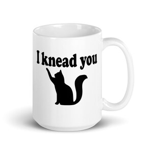 15 ounce white ceramic coffee mug with a pun image of a silhouette of a cat and the words I knead you printed on both sides of the mug.