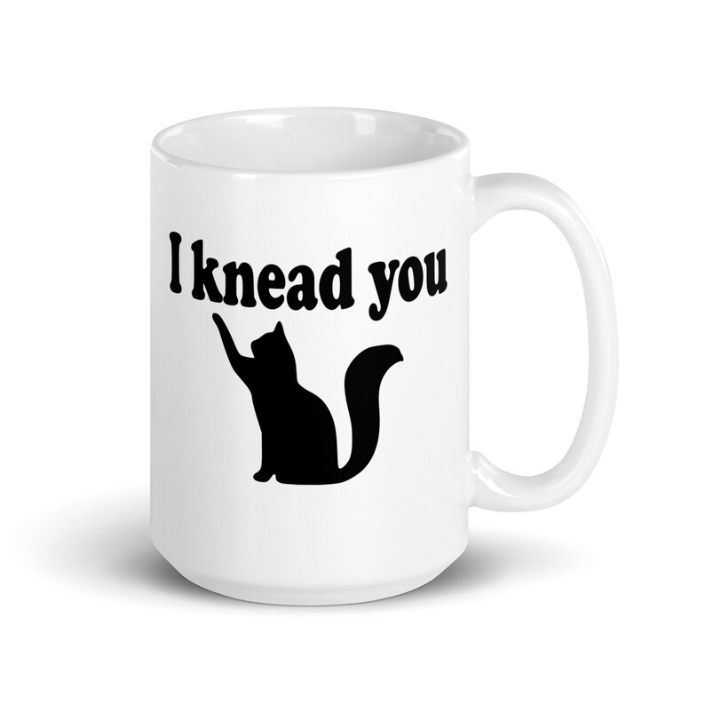 15 ounce white ceramic coffee mug with a pun image of a silhouette of a cat and the words I knead you printed on both sides of the mug.