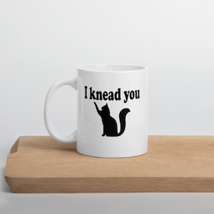 20 ounce white ceramic coffee mug with a pun image of a silhouette of a cat and the words I knead you printed on both sides of the mug.