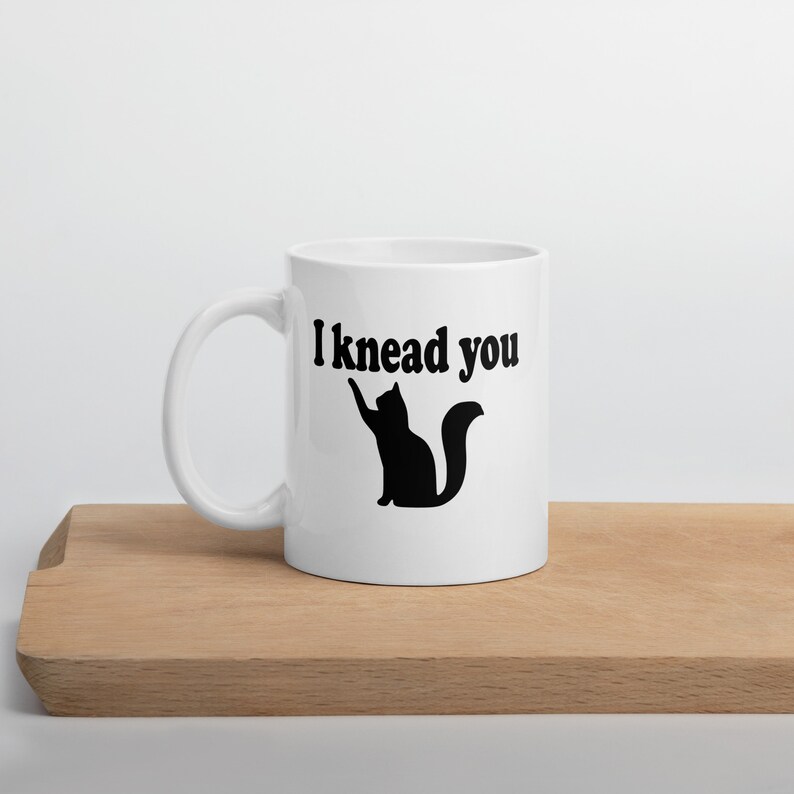 20 ounce white ceramic coffee mug with a pun image of a silhouette of a cat and the words I knead you printed on both sides of the mug.