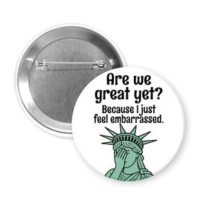 May include: A white button with the text "Are we great yet? Because I just feel embarrassed." features a green illustration of the Statue of Liberty covering her eyes. The button has a silver pin back.