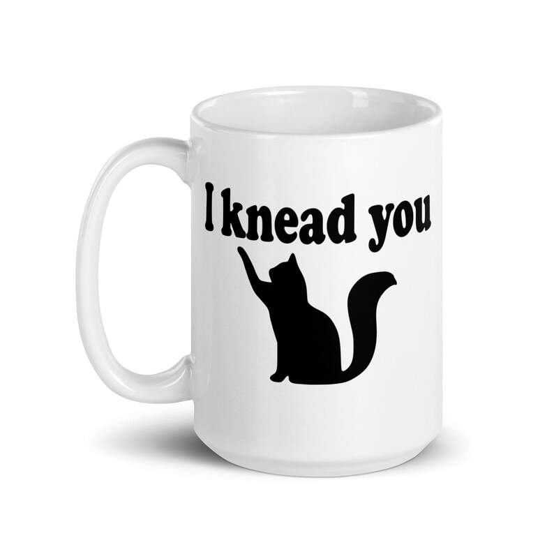 15 ounce white ceramic coffee mug with a pun image of a silhouette of a cat and the words I knead you printed on both sides of the mug.