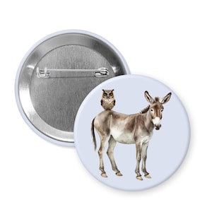 May include: A round button pin featuring a watercolor illustration of a donkey with an owl perched on its back. The donkey is brown and gray, and the owl is brown. The pin has a light blue background.