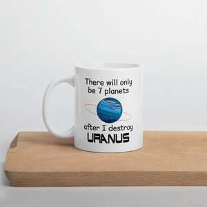 May include: White ceramic mug with a handle, featuring the text "There will only be 7 planets after I destroy URANUS." The design includes a blue planet with a red ring. The mug is placed on a wooden surface.