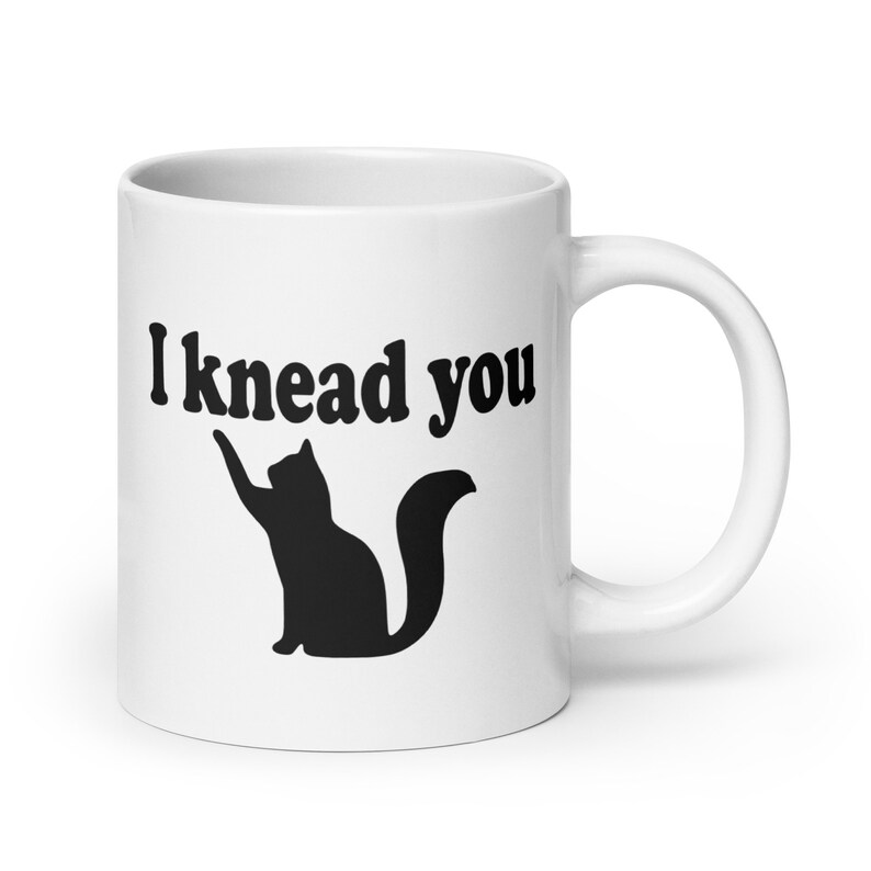 20 ounce white ceramic coffee mug with a pun image of a silhouette of a cat and the words I knead you printed on both sides of the mug.