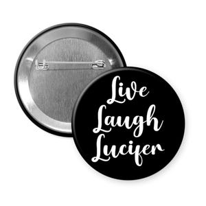 May include: A black button with the words "Live Laugh Lucifer" in white cursive script. The button has a metal pin back and a shiny silver finish. The button is round and has a simple, bold design.