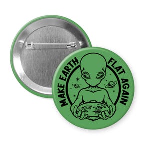 May include: A green button with a silver pin back. The button features a black illustration of an alien holding a flat Earth, with the text "MAKE EARTH FLAT AGAIN" in a circular design.