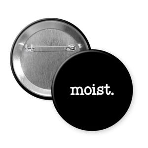 May include: A black button with the word "moist" in white, sans-serif font. The button is round and has a pin on the back. Another button is partially visible, showing the pin backing.