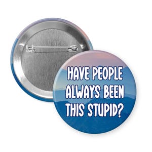 May include: Two round badges with a mountain landscape design. One badge shows the back with a pin. The other badge has the text "HAVE PEOPLE ALWAYS BEEN THIS STUPID?" in white letters.