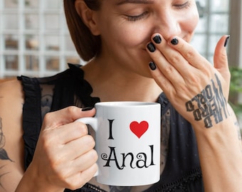 Op de afbeelding: Witte keramische mok met de woorden "I ❤️ Anal" in zwarte tekst en een rood hart. De mok heeft een wit handvat. De mok wordt door een persoon vastgehouden.