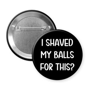 May include: A black button with the text "I SHAVED MY BALLS FOR THIS?" in white. The button is round and has a metal pin back. Another button is visible in the background.