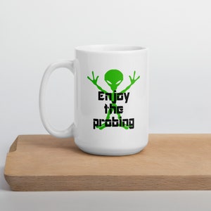 May include: White ceramic coffee mug featuring a green alien graphic and the text "Enjoy the Probing".