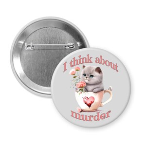 May include: A round button with a gray background and a cartoon image of a gray kitten in a teacup with pink flowers. The text "I think about murder" is written around the image in pink.