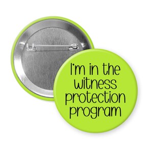 May include: A round lime green pin-back button featuring the black text "I'm in the witness protection program" printed in a simple sans-serif font. The back of the button is metallic silver with a horizontal pin mechanism for attachment.