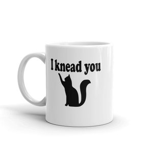 11 ounce white ceramic coffee mug with a pun image of a silhouette of a cat and the words I knead you printed on both sides of the mug.