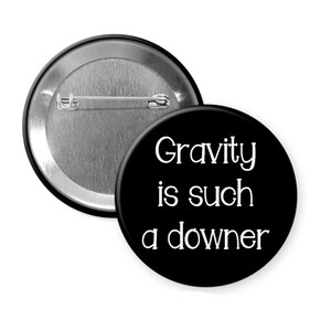May include: Two round black buttons with a metal pin back. One button displays the text "Gravity is such a downer" in white, sans-serif font. The other button is silver.