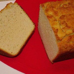 May include: A loaf of golden-brown bread, with a slice cut and placed beside the loaf. The bread sits on a red surface, with a white plate in the background. The bread has a textured crust and a soft interior.
