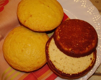 May include: Three golden-yellow, round baked goods, possibly muffins or biscuits, are arranged on a white plate. One is cut open, revealing a light interior. The baked goods rest on a colorful striped cloth.