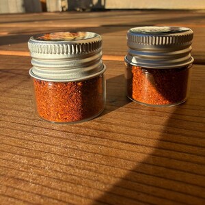 Pu&ograve; includere: Due piccoli barattoli di vetro trasparente riempiti con una spezia finemente macinata di un vibrante colore rosso-arancio. Ogni barattolo &egrave; dotato di un tappo a vite in metallo argentato e zigrinato. I barattoli sono posizionati fianco a fianco.