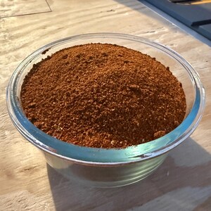 Pu&ograve; includere: Una ciotola di vetro trasparente riempita fino all'orlo con una miscela di spezie finemente macinate di un colore bruno-rossastro intenso. Il composto ha una consistenza granulosa con piccole particelle visibili di varie tonalit&agrave; di marrone e rosso.