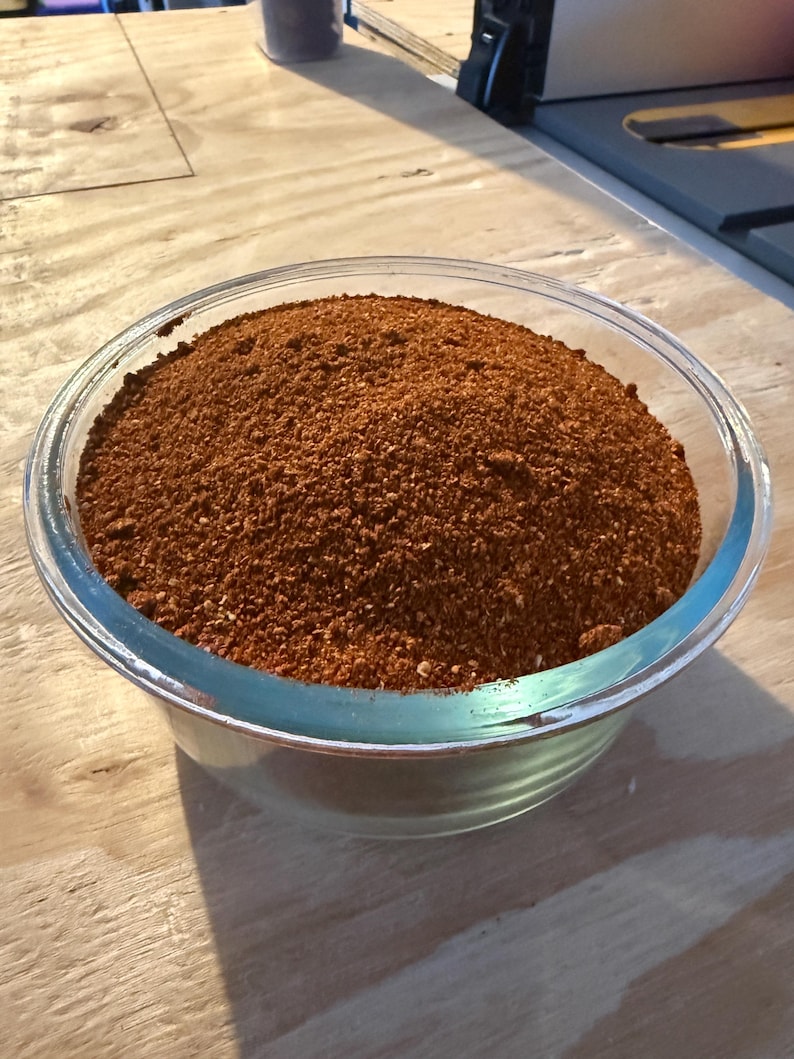 Pu&ograve; includere: Una ciotola di vetro trasparente riempita fino all'orlo con una miscela di spezie finemente macinate di un colore bruno-rossastro intenso. Il composto ha una consistenza granulosa con piccole particelle visibili di varie tonalit&agrave; di marrone e rosso.
