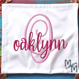 May include: White fabric square with pink and red embroidered lettering that reads "oaklynn" with a pink swirl design. The fabric is on a colorful wooden background.