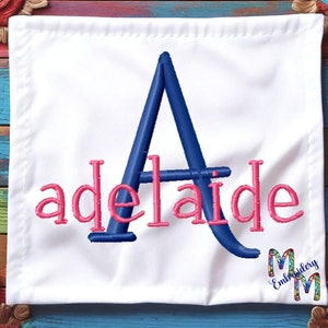 May include: White fabric square with a blue embroidered letter A and the word "adelaide" in pink embroidery. The fabric is on a colorful wooden background.