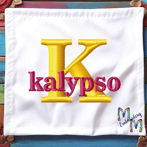 Puede incluir: Un cuadrado de tela blanca con una gran letra K bordada en amarillo y la palabra "kalypso" bordada en rosa debajo. La tela está sobre un fondo de madera de colores.