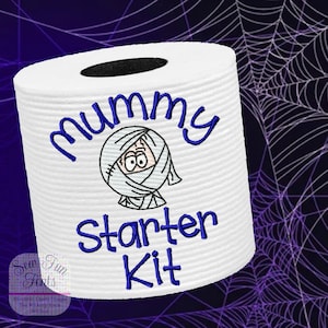 May include: A white toilet paper roll with a blue embroidered design that says "Mummy Starter Kit". The design includes a cartoon mummy with a bandage on its head.