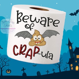 May include: A white toilet paper roll with a brown and gray bat with fangs and googly eyes. The text "Beware of CRAP-ula" is embroidered on the roll. The background is a blue sky with a silhouette of a haunted house and a graveyard.