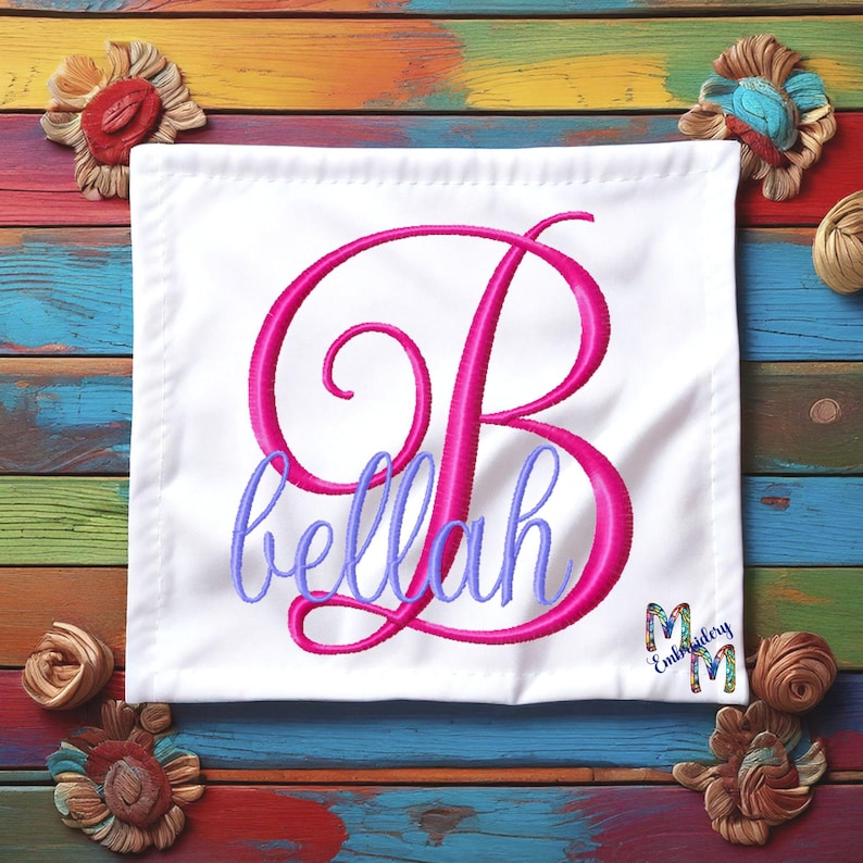 Puede incluir: Un cuadrado de tela blanca con una letra "B" bordada en rosa y el nombre "bellah" en bordado morado. La tela est&aacute; sobre un fondo de madera de colores.