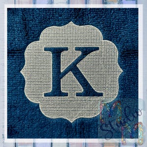 May include: Embroidered monogram letter K in a silver-coloured frame on a dark blue background.