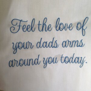 Puede incluir: Tela blanca con texto bordado en azul que dice "Siente el amor de los brazos de tu pap&aacute; alrededor tuyo hoy."